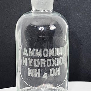 May include: Clear glass bottle with a ground glass stopper. The bottle has a rounded shape and features the embossed text "AMMONIUM HYDROXIDE NH₄OH" within an oval frame. The bottle is empty.