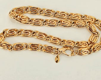Puede incluir: Un collar de cadena en tono dorado que presenta una serie de eslabones repetitivos entrelazados en forma de S. El collar incluye un cierre de mosquetón y una pequeña etiqueta metálica de forma ovalada que cuelga cerca del cierre.