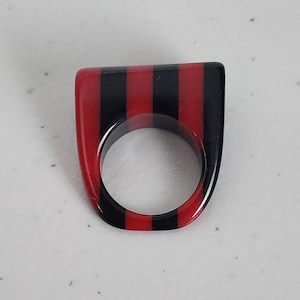 May include: A bold, rectangular ring with a flat top. The ring features alternating vertical stripes of red and black. The ring is made of a glossy material and has a circular opening for the finger.