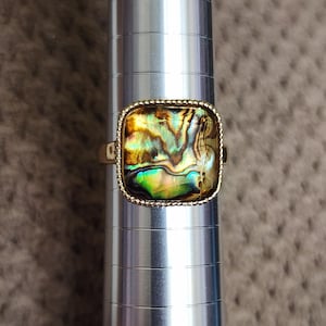May include: A gold-toned ring with a square abalone shell center. The iridescent shell displays shades of green, blue, and gold. The ring is set on a silver ring sizer. The ring's setting has a beaded border.