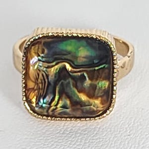 May include: A gold-toned ring with a square, iridescent abalone shell center. The ring has a textured band and a decorative border around the abalone. The abalone shell displays a mix of green, gold, and brown colors.