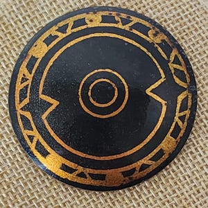 May include: A round, black decorative object with a gold-colored geometric design. The design includes a central circle, an outer ring, and angular shapes. The object has a slightly aged appearance.