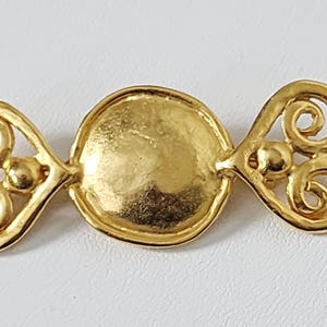 May include: A gold-colored brooch with a central circular design and ornate, swirling details on each end. The brooch has a polished finish and is set against a white background. The design is symmetrical and elegant.