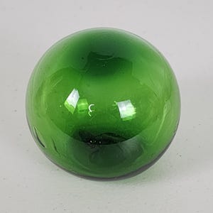 May include: A round, transparent, emerald green glass ball. The sphere reflects light, creating bright highlights. The glass has a smooth surface. The object is a decorative item.