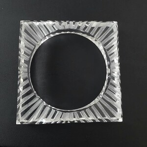 Puede incluir: Un posavasos cuadrado de cristal transparente con un recorte circular en el centro y una serie de ranuras lineales grabadas que irradian desde el c&iacute;rculo interior hasta los bordes cuadrados, creando un patr&oacute;n texturizado.