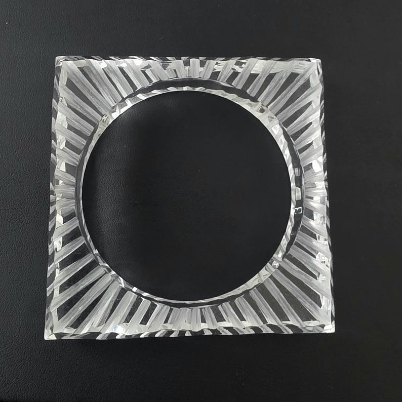 Puede incluir: Un posavasos cuadrado de cristal transparente con un recorte circular en el centro y una serie de ranuras lineales grabadas que irradian desde el c&iacute;rculo interior hasta los bordes cuadrados, creando un patr&oacute;n texturizado.