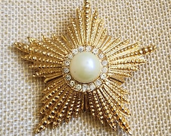 May include: Gold-toned starburst brooch with a central faux pearl surrounded by clear rhinestones. The starburst design features radiating rays and a textured surface, creating a vintage-inspired accessory.