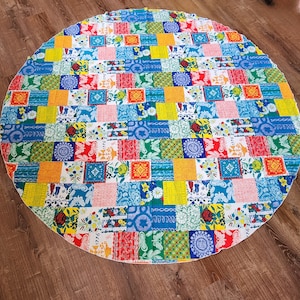 May include: A round tablecloth with a patchwork design featuring colorful squares and floral patterns. The tablecloth is made of a white fabric with a variety of colors, including red, blue, yellow, green, and orange.