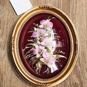 May include: Oval framed floral art piece. The artwork features a bouquet of white and lavender flowers with gold-colored leaves, set against a deep red background. The frame is gold with ornate detailing.