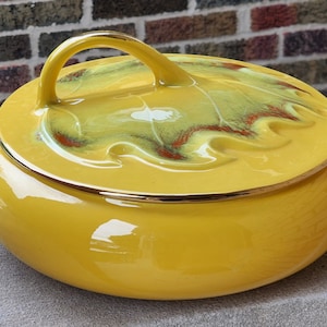 May include: A bright yellow ceramic serving dish with a lid. The lid features a leaf design with green, red, and white accents. The dish has a gold trim and a handle on the lid. It also has two side handles.