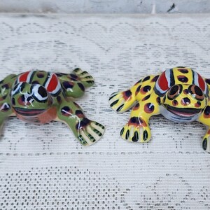 May include: Two ceramic frog figurines, one green with red and black spots, the other yellow with red and black spots. Both frogs have black eyes and a smiling mouth.