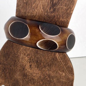 May include: A brown wooden bangle bracelet with four oval-shaped cutouts. The bracelet is a rich, dark brown color with lighter brown detailing within the cutouts. The bracelet is displayed on a wooden stand.