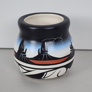 May include: A small, round ceramic pot with a black exterior and a white interior. The pot features a landscape design with black, white, blue, and orange elements. The design includes stylized mountains and geometric patterns.