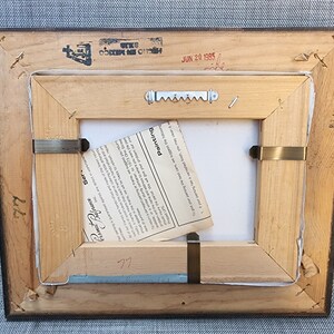 May include: The back of a wooden picture frame with a white label that reads "Painting", "Ser", "Preference", and "77". The frame has a metal hanging hook and a small piece of paper with the text "Burnished" on it. The frame also has a stamp that reads "HECHO EN MEXICO" and "JUN 20 1985".