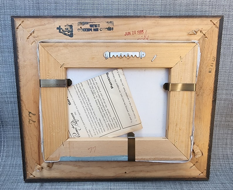 May include: The back of a wooden picture frame with a white label that reads "Painting", "Ser", "Preference", and "77". The frame has a metal hanging hook and a small piece of paper with the text "Burnished" on it. The frame also has a stamp that reads "HECHO EN MEXICO" and "JUN 20 1985".