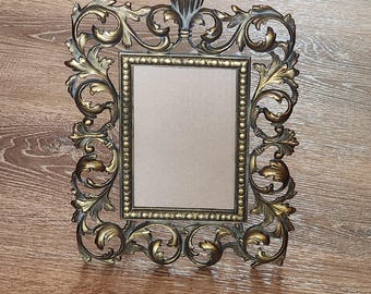 May include: A gold-toned metal picture frame with an ornate, scrolled design. The frame has a rectangular opening for a photo or artwork.