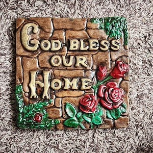May include: A square, decorative plaque with the words "God Bless Our Home" in gold lettering. The plaque has a brick-like background with red roses and green leaves.