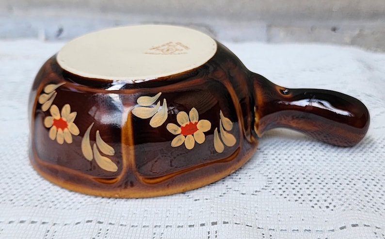 Op de afbeelding: Een bruine keramische serveerschaal met een handvat. De schaal is versierd met witte bloemen met rode centra en gouden bladeren.
