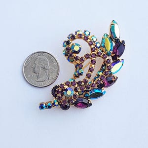 May include: A gold-toned brooch with a swirling design, adorned with purple, teal, and iridescent rhinestones. The brooch is approximately 2 inches in length and features a pin closure on the back. A quarter is next to the brooch for size comparison.