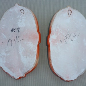 May include: Two white ceramic plaques with orange trim and a hanging loop. The plaque on the left has the number 27 and the number 171 written on it. The plaque on the right has the number 44 and the number 147 written on it.