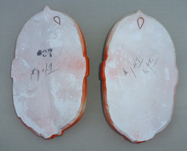 May include: Two white ceramic plaques with orange trim and a hanging loop. The plaque on the left has the number 27 and the number 171 written on it. The plaque on the right has the number 44 and the number 147 written on it.