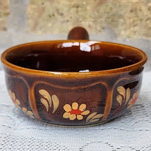 Op de afbeelding: Een bruine keramische kom met een houten handvat. De kom is versierd met gele en oranje bloemen en bladeren.