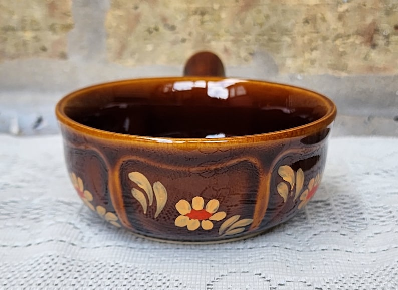 Op de afbeelding: Een bruine keramische kom met een houten handvat. De kom is versierd met gele en oranje bloemen en bladeren.