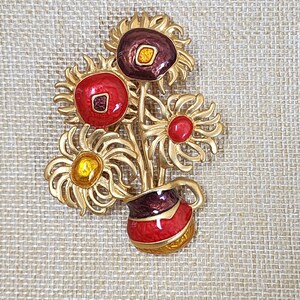 May include: A gold-tone brooch featuring a bouquet of stylized sunflowers with red, yellow, and purple enamel accents. The sunflowers are arranged in a vase with red and purple enamel.
