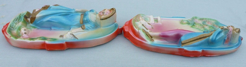 May include: Two ceramic wall plaques depicting women in a reclining pose. The plaques are painted in a pastel color scheme with blue, pink, and green accents. The women are wearing dresses and have long hair.