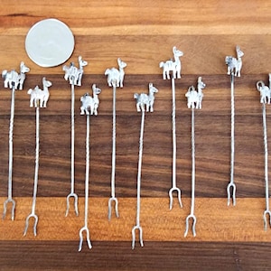 May include: A collection of silver-colored cocktail stirrers with llama-shaped toppers. The stirrers have a twisted stem design and a small fork at the bottom. A small, round, silver-colored object is also present. The items are arranged on a wooden surface.