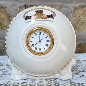 May include: Cream-colored ceramic clock with a gold-tone clock face and Roman numerals. The clock features a Claddagh design with the words "A Symbol of Love & Friendship". The clock has a decorative gold trim.