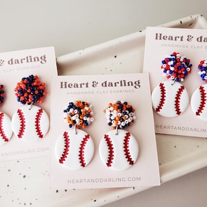 May include: Handmade clay earrings featuring a baseball design. The earrings have a white baseball with red stitching and a cluster of small beads in red, white, and blue or orange and navy blue. The text "Heart & darling" is printed on the cards.