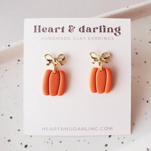 May include: A pair of orange clay pumpkin earrings with gold-tone metal bows attached to the top. The earrings are displayed on a white card printed with the text Heart & darling HANDMADE CLAY EARRINGS and the website address HEARTANDDARLING.COM.