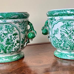 May include: Two white ceramic planters with green floral designs and handles. The planters have a rounded base and a wide opening.