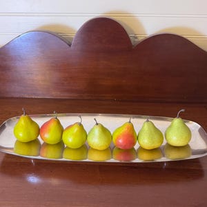 May include: A long, oval-shaped silver-toned metal tray holding seven fresh pears. The pears are primarily yellow-green with some red blushing on their skins. Each pear features a thin brown stem at the top, and they are arranged in a single row.