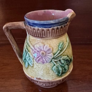 May include: A ceramic creamer pitcher featuring a raised floral design with a purple flower and green leaves on a yellow background. The pitcher has a blue rim, a brown handle, and a basket-weave texture at the base with a pink interior finish.