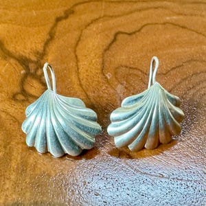 May include: A pair of silver-toned earrings shaped like fan-style seashells. Each earring features a curved, ribbed surface with a metallic finish and a simple hook-style wire attachment at the top for hanging. The design is symmetrical and smooth.