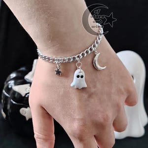 Peut inclure: Un bracelet à maillons gourmette argenté orné de trois breloques pendantes : une petite étoile à cinq branches, un fantôme blanc aux yeux noirs et un croissant de lune. Le bracelet comporte un logo avec le texte of stars and wine.