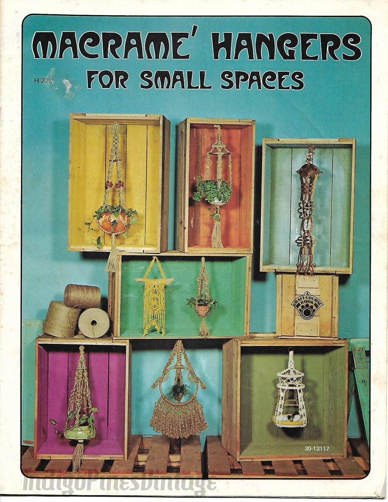 Może przedstawiać: Okładka książki z tytułem "Macrame' Hangers For Small Spaces" w pogrubionych, czarnych literach. Okładka przedstawia kolaż makramowych wieszak&oacute;w na rośliny prezentowanych w drewnianych skrzyniach.