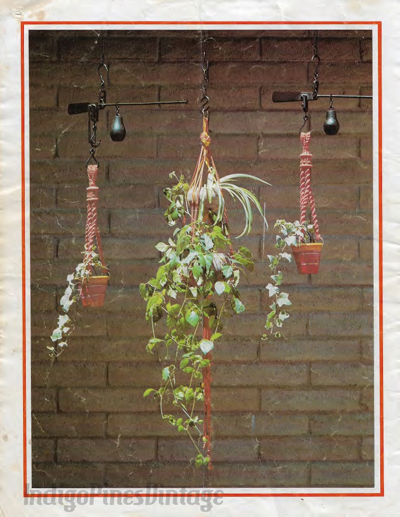 May include: Three hanging plant pots made of macrame cord, with a brick wall background. The pots are suspended from a metal scale with a weight on each side.