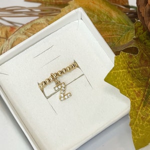 Op de afbeelding: Een goudkleurige kralenring met een hangende letter Z-bedel bezet met kleine doorzichtige steentjes. De ring wordt gepresenteerd in een vierkant wit doosje, geplaatst tegen een achtergrond van herfstkleurige bladeren en een dennenappel.