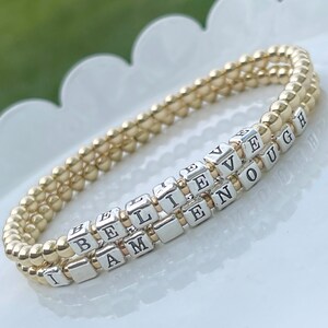 May include: A gold and silver beaded bracelet with the words "I am enough" and "Believe" spelled out in silver beads.