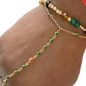 Könnte beinhalten: Ein goldenes Kettenarmband mit grünen und orangefarbenen Edelsteinen. Das Armband ist mit einem goldenen Perlenarmband mit schwarzen, bernsteinfarbenen und grünen Perlen geschichtet.