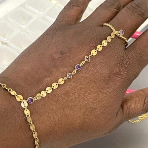 Könnte beinhalten: Eine goldene Handkette mit Ring- und Armband-Design. Die Kette hat kleine, runde goldene Scheiben und ist mit lila Edelsteinen in runden und quadratischen Fassungen verziert. Der Schmuck wird an einer Hand getragen.