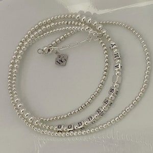 Può includere: Tre braccialetti d'argento con perline e ciondoli con lettere che compongono i nomi "GOD", "DES" e "AMY". Un piccolo ciondolo d'argento con le iniziali "WG" pende dal braccialetto superiore.