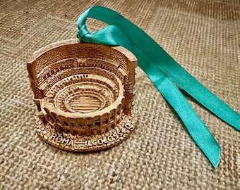 May include: A gold-colored Colosseum miniature with the text "ROMA IL COLOSSEO" at the base, attached to a teal ribbon. The detailed replica of the Roman amphitheater is a decorative item.