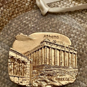 May include: A gold-colored ornament featuring a detailed relief of the Parthenon in Athens, Greece. The ornament has the word "Athens" at the top and "GREECE" at the bottom. A clear plastic backing and a white ribbon are attached for hanging.