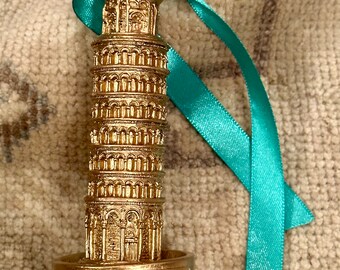May include: A gold-colored miniature replica of the Leaning Tower of Pisa, with the words "TORRE DI PISA" at the base. A teal ribbon is attached to the top. The tower is a detailed model of the famous Italian landmark.