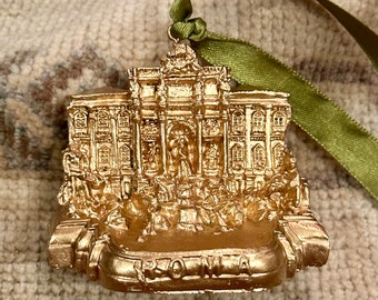 May include: A gold-colored ornament featuring a detailed replica of the Trevi Fountain in Rome. The ornament has a green ribbon for hanging and the word "ROMA" is visible at the base. The ornament is likely made of resin or a similar material.