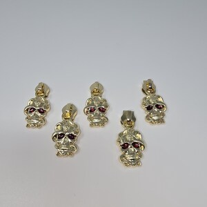 May include: Five gold-colored zipper pulls with a skull design. The skulls have red eyes and are detailed with intricate carvings.